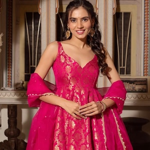 May include: A vibrant fuchsia-colored dress with gold floral brocade detailing. The sleeveless dress features a V-neckline and a flowing skirt. A matching sheer scarf with gold trim completes the ensemble. The woman is wearing gold earrings and a bracelet.