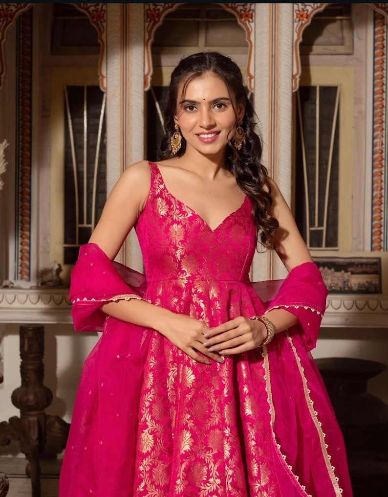 May include: A vibrant fuchsia-colored dress with gold floral brocade detailing. The sleeveless dress features a V-neckline and a flowing skirt. A matching sheer scarf with gold trim completes the ensemble. The woman is wearing gold earrings and a bracelet.