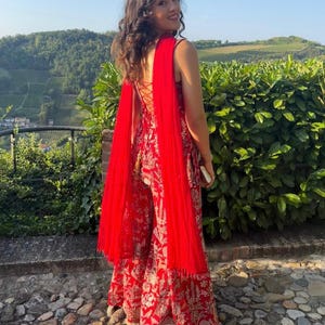 May include: A woman wearing a red and gold patterned jumpsuit with a matching red shawl. The jumpsuit has a lace-up back and wide legs. The shawl drapes over the shoulders. The background features green foliage and a stone pathway.