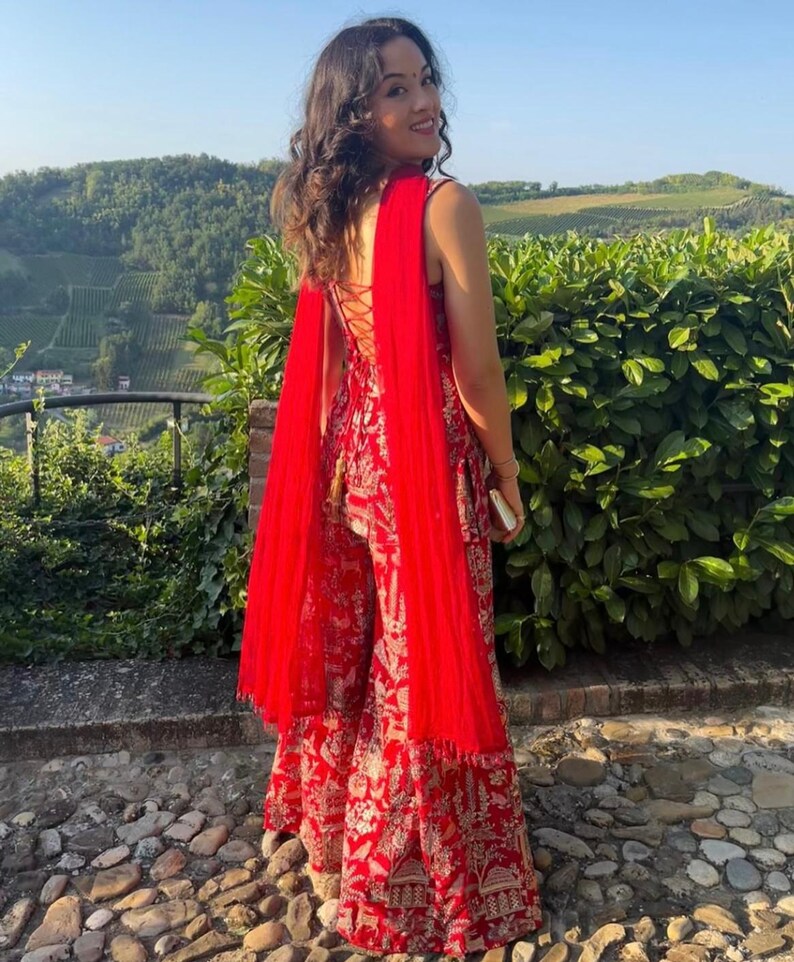 May include: A woman wearing a red and gold patterned jumpsuit with a matching red shawl. The jumpsuit has a lace-up back and wide legs. The shawl drapes over the shoulders. The background features green foliage and a stone pathway.