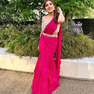 以下が含まれることがあります： ピンクのフリル付きサリーを着た女性。シルバービーズのベルトと、白いビーズのワンショルダーブラウスを着用している。