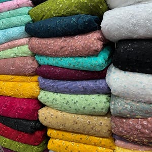 May include: A stack of fabric rolls in various colors, including yellow, green, pink, and blue. The fabric appears to be embroidered with floral patterns and sequins, suitable for apparel or craft projects.