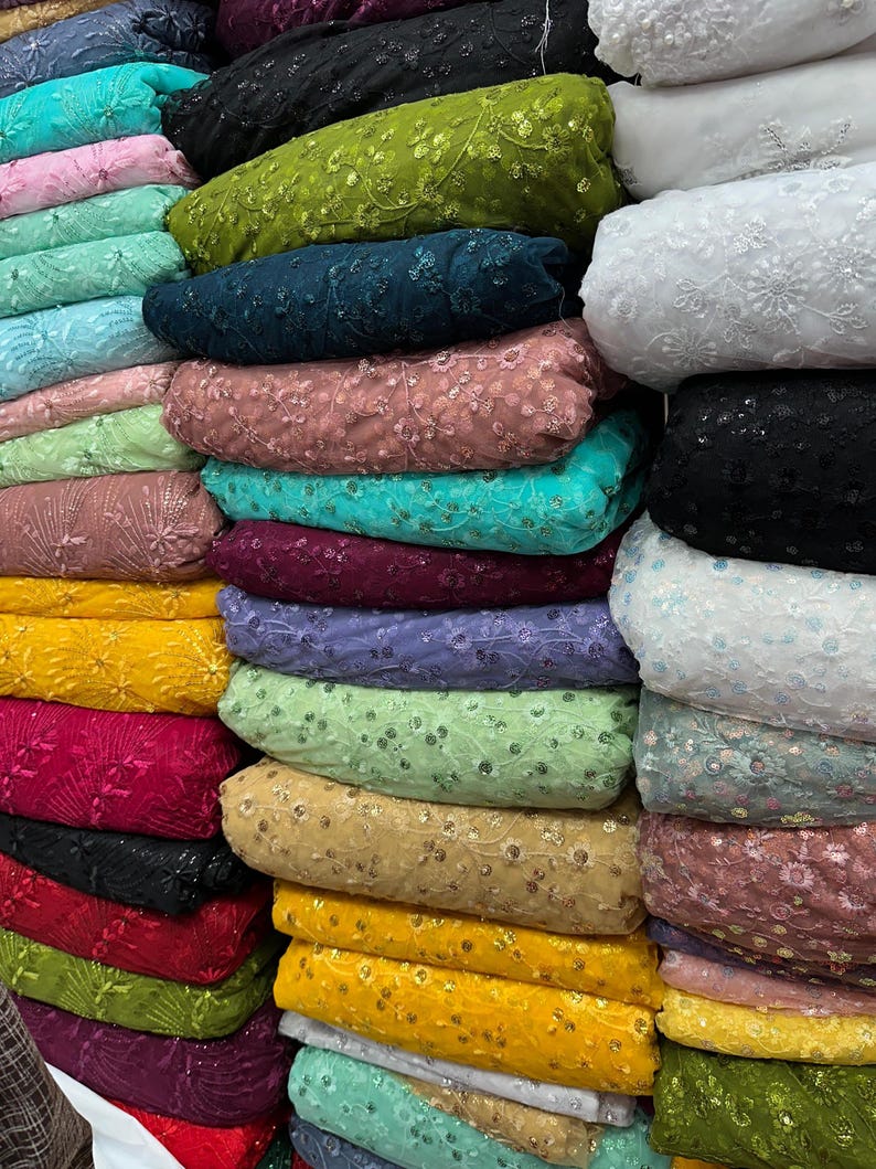 May include: A stack of fabric rolls in various colors, including yellow, green, pink, and blue. The fabric appears to be embroidered with floral patterns and sequins, suitable for apparel or craft projects.