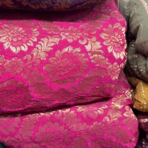 May include: Close-up of stacked fabric rolls featuring a vibrant pink and gold floral brocade pattern. The fabric has a luxurious sheen and a detailed design. Other colors visible are purple, blue, and green.