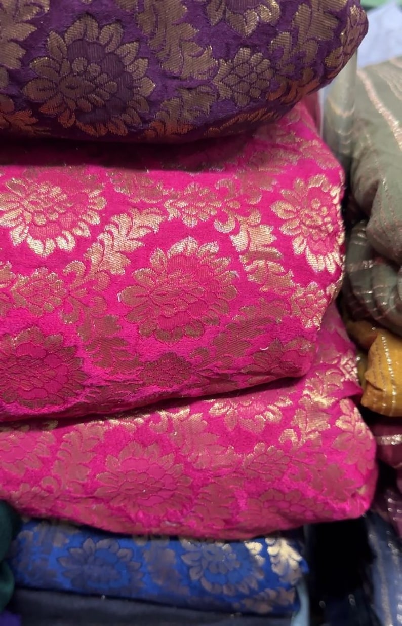 May include: Close-up of stacked fabric rolls featuring a vibrant pink and gold floral brocade pattern. The fabric has a luxurious sheen and a detailed design. Other colors visible are purple, blue, and green.