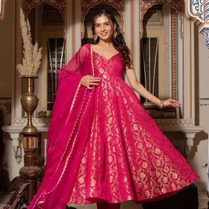 May include: A vibrant fuchsia-colored dress with gold floral patterns. The dress features a V-neckline and a flowing skirt. A matching sheer scarf with a delicate trim is draped over the shoulders. The outfit is a traditional style.
