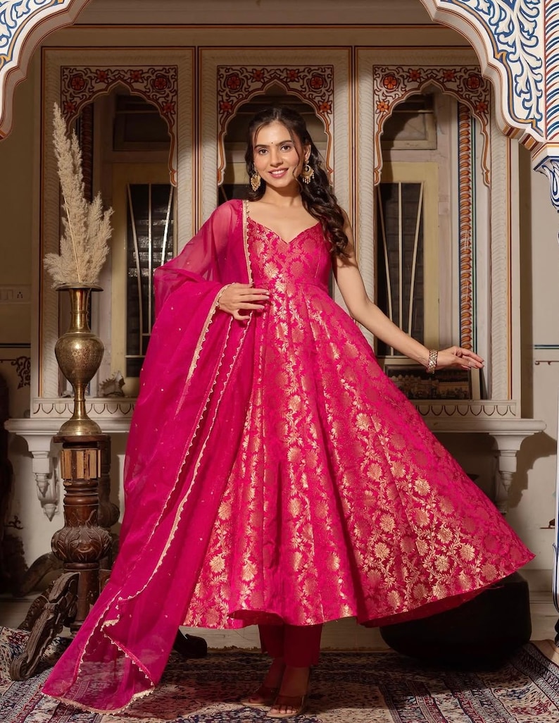 May include: A vibrant fuchsia-colored dress with gold floral patterns. The dress features a V-neckline and a flowing skirt. A matching sheer scarf with a delicate trim is draped over the shoulders. The outfit is a traditional style.