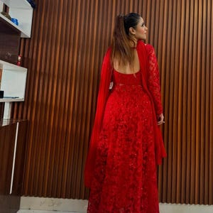 May include: A long, red, formal dress with intricate floral lace detailing. The dress features long sleeves and a matching shawl. The back of the dress has a low, open design. The background is a wooden wall.
