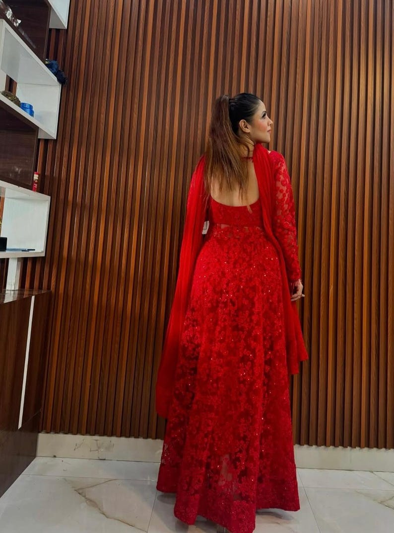 May include: A long, red, formal dress with intricate floral lace detailing. The dress features long sleeves and a matching shawl. The back of the dress has a low, open design. The background is a wooden wall.