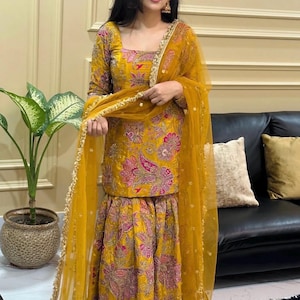 May include: Mustard yellow embroidered ensemble featuring a square-neck top, flared pants, and a sheer dupatta. The floral embroidery includes pink and green accents. Gold trim adorns the edges of the outfit and dupatta. The outfit is worn with gold earrings.