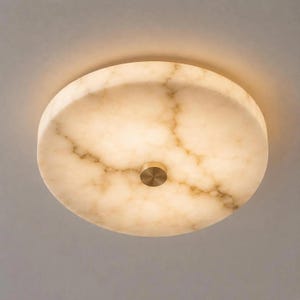 May include: A circular flush mount ceiling light fixture featuring a translucent, cream-colored stone shade with natural brown veining. The center of the fixture is secured by a small, circular, brushed metallic gold-toned metal cap.