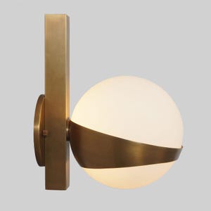 May include: A brass wall sconce with a white globe and a curved brass shade. The sconce has a rectangular brass base.