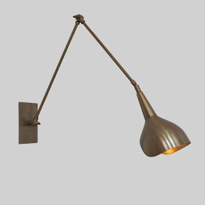 May include: A brass wall-mounted light fixture with a long, adjustable arm and a cone-shaped shade. The light is turned on and illuminating the room.