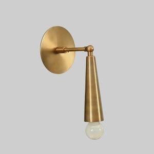May include: A brass wall sconce with a circular backplate and a conical shade. The sconce has a straight arm connecting the backplate to the shade, and a clear, round light bulb is visible at the bottom of the shade.