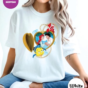 May include: White t-shirt featuring a heart-shaped locket design with Ariel and Prince Eric from The Little Mermaid. The locket is surrounded by a gold chain and includes Flounder and Sebastian. The shirt has the text "Next Day Shipping" and "White".