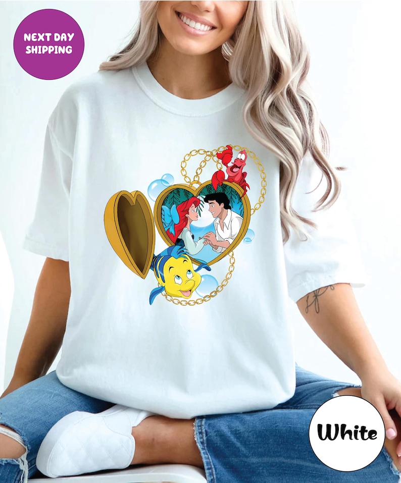 May include: White t-shirt featuring a heart-shaped locket design with Ariel and Prince Eric from The Little Mermaid. The locket is surrounded by a gold chain and includes Flounder and Sebastian. The shirt has the text "Next Day Shipping" and "White".