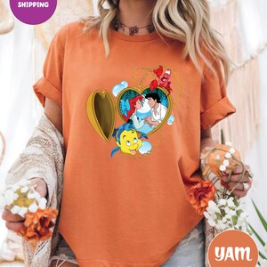 May include: Orange t-shirt featuring a heart-shaped locket with a cartoon image of Ariel and Prince Eric. The locket is surrounded by Flounder and Sebastian. The shirt has the text "Next Day Shipping" on a purple circle.