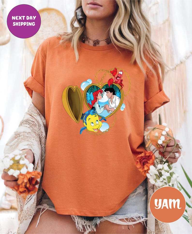 May include: Orange t-shirt featuring a heart-shaped locket with a cartoon image of Ariel and Prince Eric. The locket is surrounded by Flounder and Sebastian. The shirt has the text "Next Day Shipping" on a purple circle.
