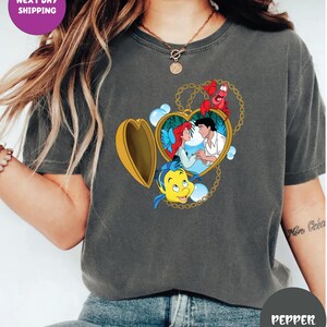 May include: Dark gray t-shirt featuring a heart-shaped locket with a Disney's The Little Mermaid design. The locket contains Ariel and Prince Eric, with Flounder and Sebastian. The shirt also has the text "NEXT DAY SHIPPING".