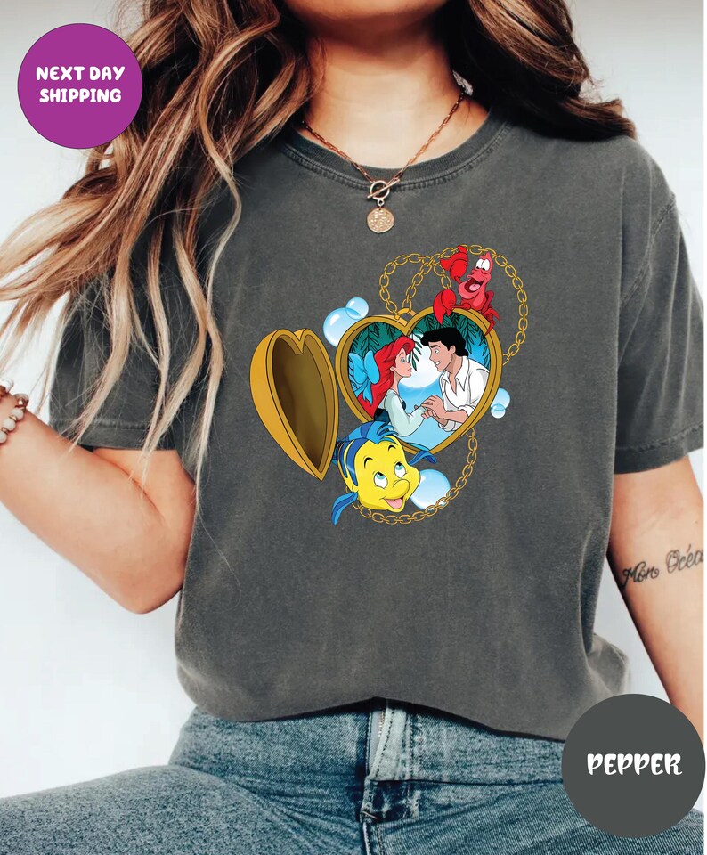 May include: Dark gray t-shirt featuring a heart-shaped locket with a Disney's The Little Mermaid design. The locket contains Ariel and Prince Eric, with Flounder and Sebastian. The shirt also has the text "NEXT DAY SHIPPING".