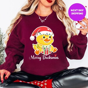 May include: Burgundy sweatshirt featuring a cartoon yellow duck wearing a Santa hat and holding a wrapped gift. The duck is adorned with Christmas lights. The text "Merry Ducksmas" is written below the duck in white script.