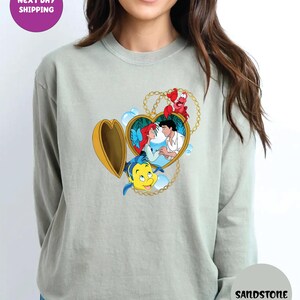 May include: A light green sweatshirt featuring a heart-shaped locket with a cartoon image of Ariel and Prince Eric. The locket is gold with a gold chain. The sweatshirt has the word "Sandstone" printed on it.