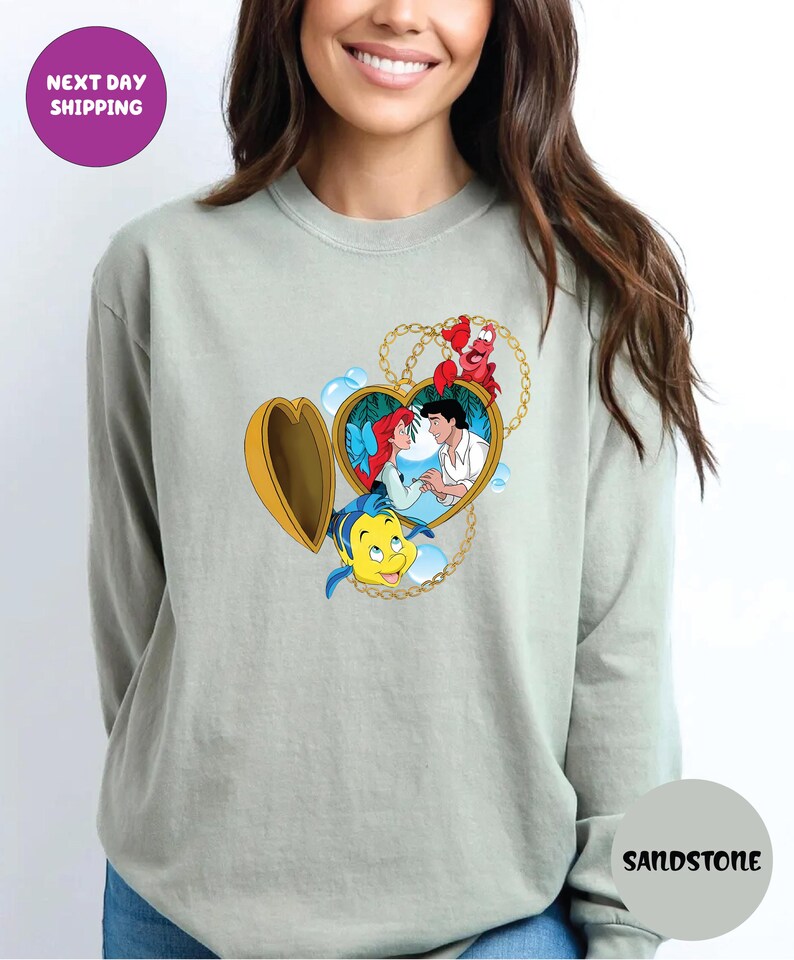 May include: A light green sweatshirt featuring a heart-shaped locket with a cartoon image of Ariel and Prince Eric. The locket is gold with a gold chain. The sweatshirt has the word "Sandstone" printed on it.