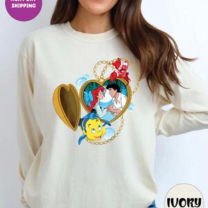 May include: Ivory long-sleeve sweatshirt featuring a heart-shaped locket design with characters from The Little Mermaid. The locket is gold with a chain, and the characters include Ariel, Prince Eric, Flounder, and Sebastian. The text "NEXT DAY SHIPPING" is in a purple circle.