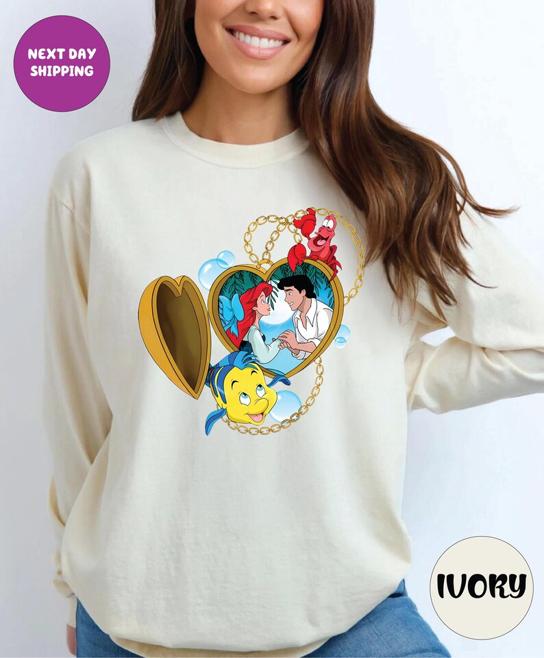 May include: Ivory long-sleeve sweatshirt featuring a heart-shaped locket design with characters from The Little Mermaid. The locket is gold with a chain, and the characters include Ariel, Prince Eric, Flounder, and Sebastian. The text "NEXT DAY SHIPPING" is in a purple circle.