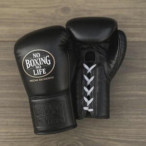 Könnte beinhalten: Paar schwarze Boxhandschuhe mit weißen Schnürsenkeln. Einer der Handschuhe hat ein rundes Logo mit dem Text "NO BOXING NO LIFE" und "HECHO EN MEXICO". Die Handschuhe sind aus Leder und für das Boxen konzipiert.