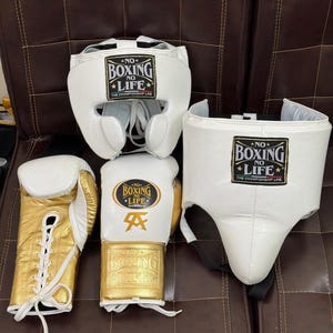 May include: A boxing set featuring a white headgear, a white groin protector, and a pair of gold and white boxing gloves. The gloves and headgear have the text "NO BOXING NO LIFE" printed on them.