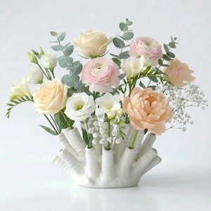 May include: A floral arrangement featuring a variety of flowers in shades of white, pink, and peach, including roses and peonies. The flowers are arranged in a white, textured vase with multiple openings, creating a unique display.