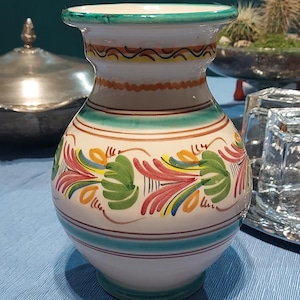 Może przedstawiać: Ręcznie malowany ceramiczny wazon z białą podstawą, ozdobiony kolorowymi wzorami kwiatowymi w kolorze zielonym, czerwonym, żółtym i niebieskim. Wazon ma zieloną obwódkę i ma około 15 cm wysokości.