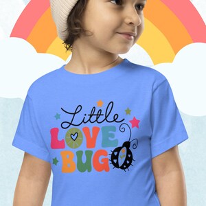 May include: A light blue t-shirt with a colorful graphic that says "Little Love Bug" and features a ladybug with a heart on its back.