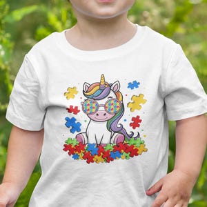 May include: A white t-shirt with a colorful cartoon unicorn wearing rainbow puzzle piece glasses. The unicorn is sitting on a bed of colorful puzzle pieces.