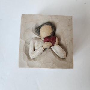 Könnte beinhalten: Eine geschnitzte Holzfigur einer Person, die ein rotes Herz hält. Die Figur trägt ein langärmeliges, cremefarbenes Kleidungsstück und ist vor einem strukturierten, beigefarbenen Hintergrund platziert. Das Herz hat eine tiefrote Farbe.