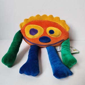 May include: A plush toy shaped like a round orange creature with a yellow scalloped top, two green arms, and two blue legs. The face features two large yellow eye patches with dark blue pupils, a dark blue nose, and a wide dark blue stitched mouth.