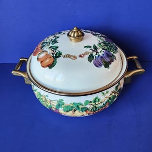 Op de afbeelding: Witte emaille ovenschaal met deksel, met een kleurrijk fruit- en bladerenontwerp. De schaal heeft goudkleurige handvatten en randen, en een goudkleurige knop op het deksel. Het ontwerp omvat perziken, druiven en pruimen.