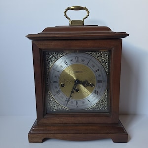 May include: A wooden mantel clock with a dark brown finish and a brass handle on top. The clock face features a gold-toned centre, a silver-toned outer ring with black Roman numerals, ornate gold corner accents, and black metal clock hands.