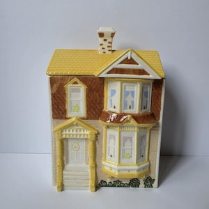 May include: A ceramic house-shaped canister featuring a yellow roof and chimney, brown textured walls, and a cream-colored lower section. The front displays a white door with a letter L, a bay window, and painted windows with blue curtains and green accents.