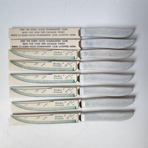 Op de afbeelding: Een set van zes vintage Quikut roestvrijstalen messen met wit gemarmerde handvatten. Elk mes zit in de originele doos, met de tekst "JOIN THE ROBIN HOOD HOMEMAKERS' CLUB!" en "Quikut STAINLESS".