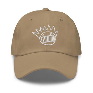 May include: A tan baseball cap with a white embroidered cartoon face design. The cap has a curved brim and an adjustable strap at the back. The embroidered design features a crown and a mouth.