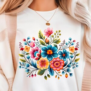 Könnte beinhalten: Ein weißes T-Shirt mit einem farbenfrohen Blumendesign. Die Blumen sind blau, rosa, gelb und grün. Das Design ist mittig auf dem Shirt platziert.