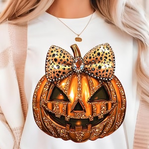 Può includere: T-shirt bianco con un design dettagliato di zucca gioiello e un fiocco. La zucca è arancione con una superficie gioiello, una faccia nera e un fiocco dorato con pois neri. È visibile una collana d'oro.