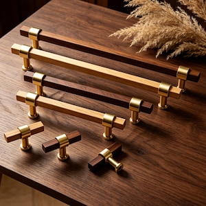 Puede incluir: Un conjunto de tiradores y pomos de madera para armarios. Los tiradores tienen un diseño rectangular con un centro de madera marrón oscuro y extremos de madera clara, acentuados con anillos y bases de metal dorado. Los pomos tienen un diseño en forma de T.