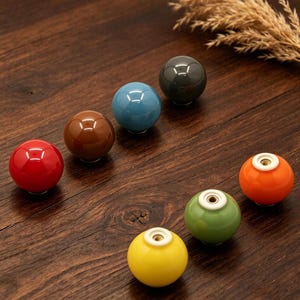May include: A collection of colorful ceramic cabinet knobs in red, brown, blue, green, yellow, orange, and dark gray. The knobs are round with a glossy finish and sit on a dark wood surface.