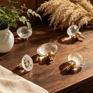 Puede incluir: Pomos de armario de vidrio transparente con bases doradas. Los pomos tienen un diseño facetado y están dispuestos sobre una superficie de madera. Los elementos decorativos incluyen un pequeño jarrón blanco con vegetación y hierba ornamental seca.