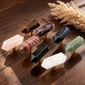 Può includere: Una collezione di otto pomelli per mobili in pietre preziose di vari colori e motivi. I pomelli sono montati su basi color oro e disposti su una superficie di legno. I colori includono rosa, marrone, viola, nero, verde e bianco.