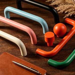 Puede incluir: Una colección de herrajes de gabinete coloridos, que incluyen manijas, perillas y una esfera. Los herrajes son de varios colores: negro, azul claro, rosa, crema, naranja y verde. Un cuaderno de cuero y un bolígrafo están en primer plano.