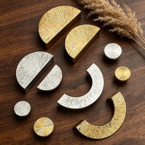 May include: An assortment of gold and silver cabinet hardware, including half-moon pulls, curved handles, and round knobs. The hardware features a textured, sunburst-like design. The pieces are arranged on a dark wood surface, with dried ornamental grass in the background.